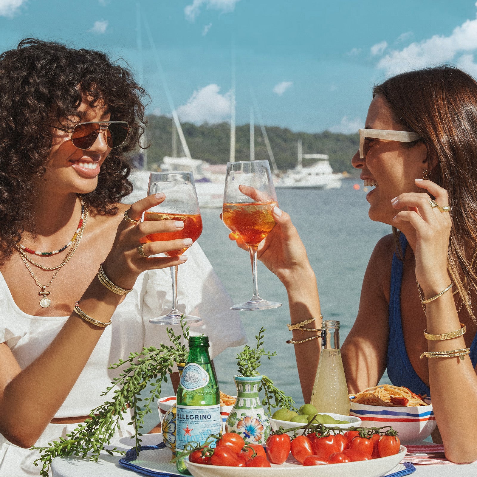 Two women outdoors with a harbor in the background, seated, raising glasses of sparkling water and toasting