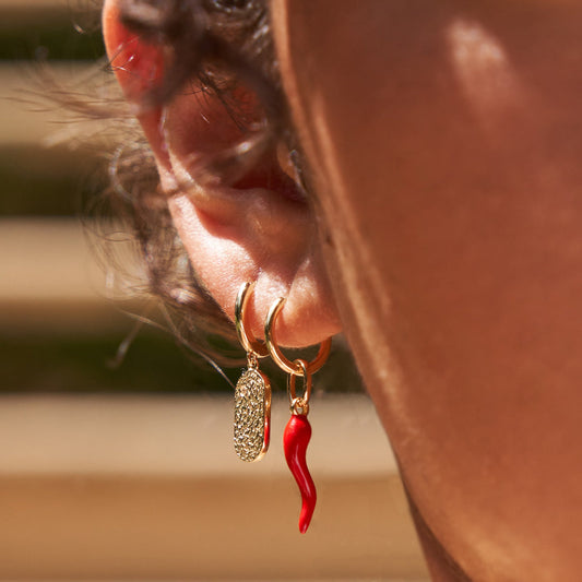 Two distinct Arms Of Eve handmade earrings, including the red Cornicello, being modeled on a woman's ear in sunlight